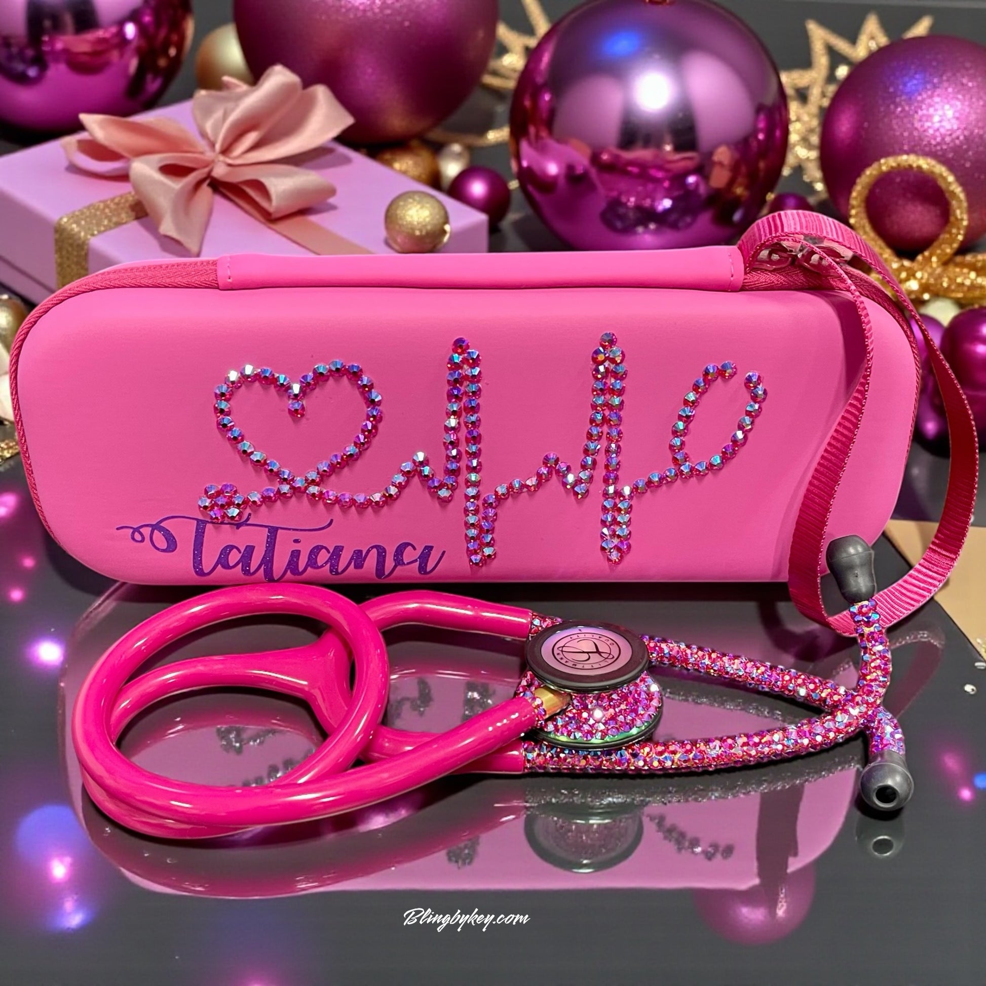Pink stethoscope with rhinestone details and matching case on a reflective surface with decorative background