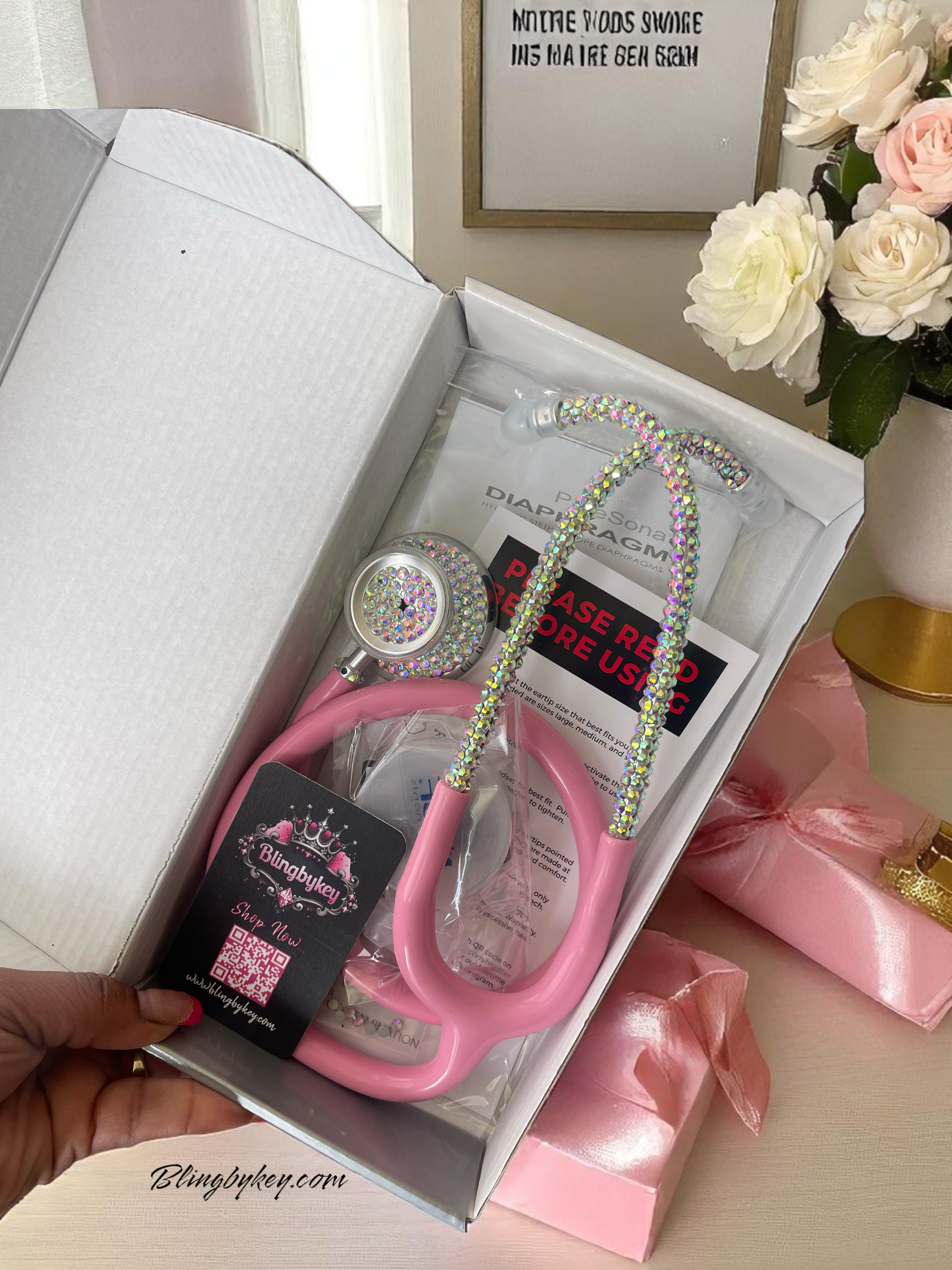 Pink stethoscope with decorative elements in a box, surrounded by flowers and ribbons.