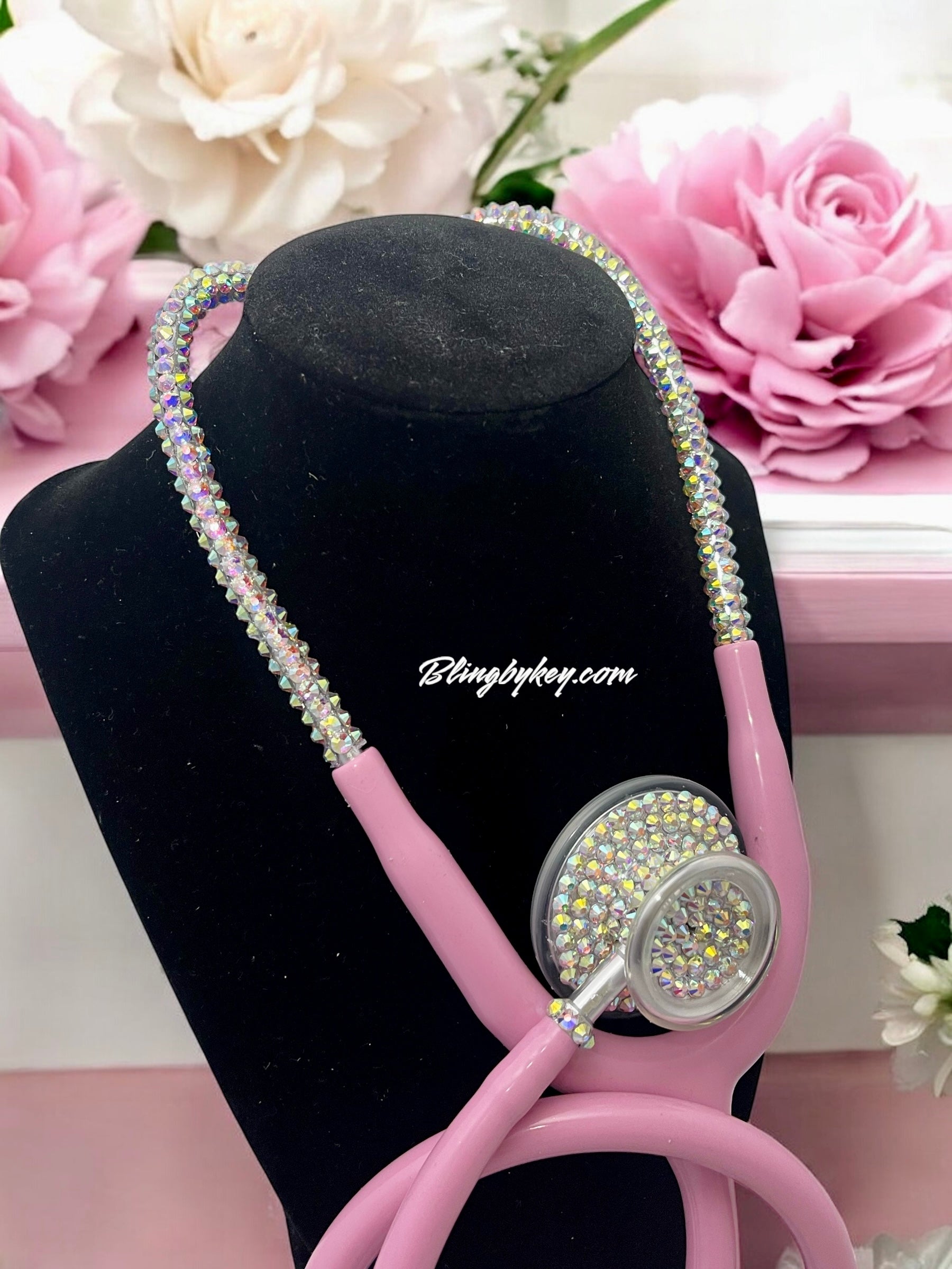 Pink stethoscope with rhinestone embellishments on a black stand with flowers in the background